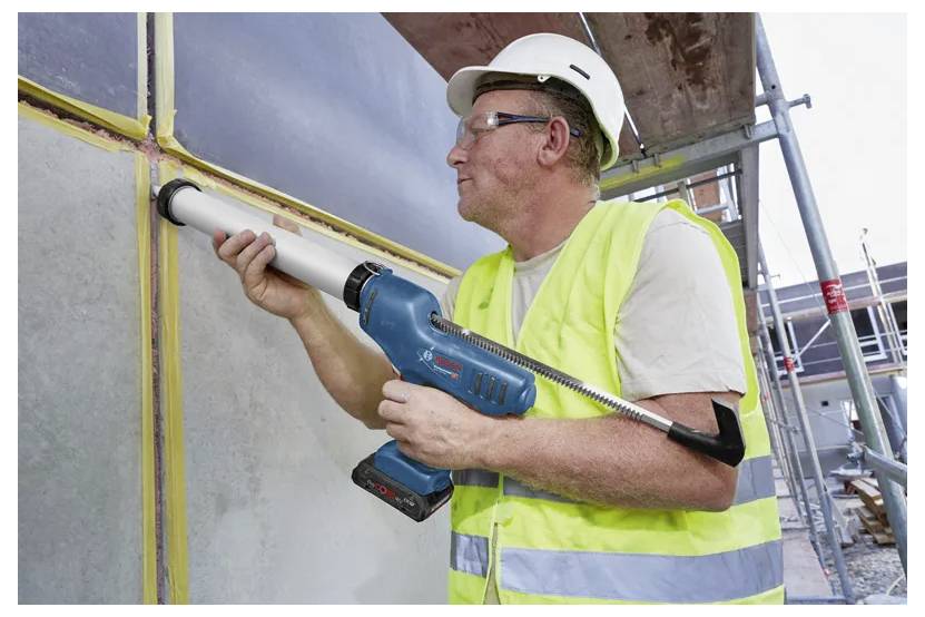 Un ouvrier du bâtiment portant un casque de chantier et un gilet de sécurité utilise un pistolet à calfeutrer électrique pour combler les interstices entre les panneaux muraux sur un chantier de construction.