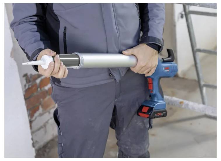 Une personne tenant un pistolet à calfeutrer sans fil, portant des vêtements de travail, debout sur un chantier de construction avec des briques apparentes et une échelle à proximité.