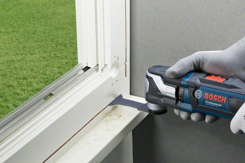 'Une main tient un outil multifonction Bosch et coupe précisément un montant de fenêtre. À l'extérieur, on aperçoit un gazon vert.'