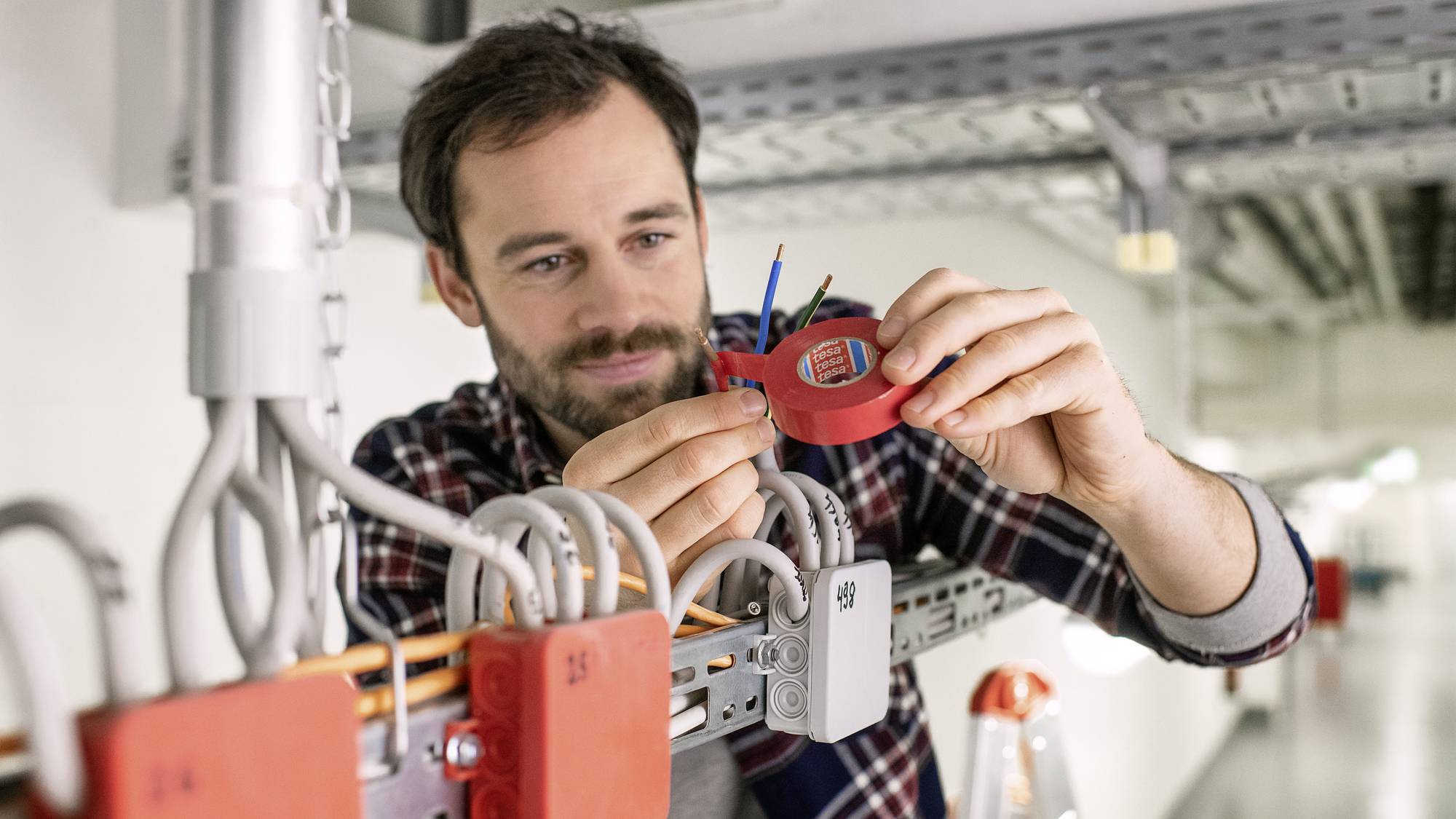 Un homme travaille sur un câblage électrique dans un environnement industriel. Il tient un appareil rouge et fixe des fils.