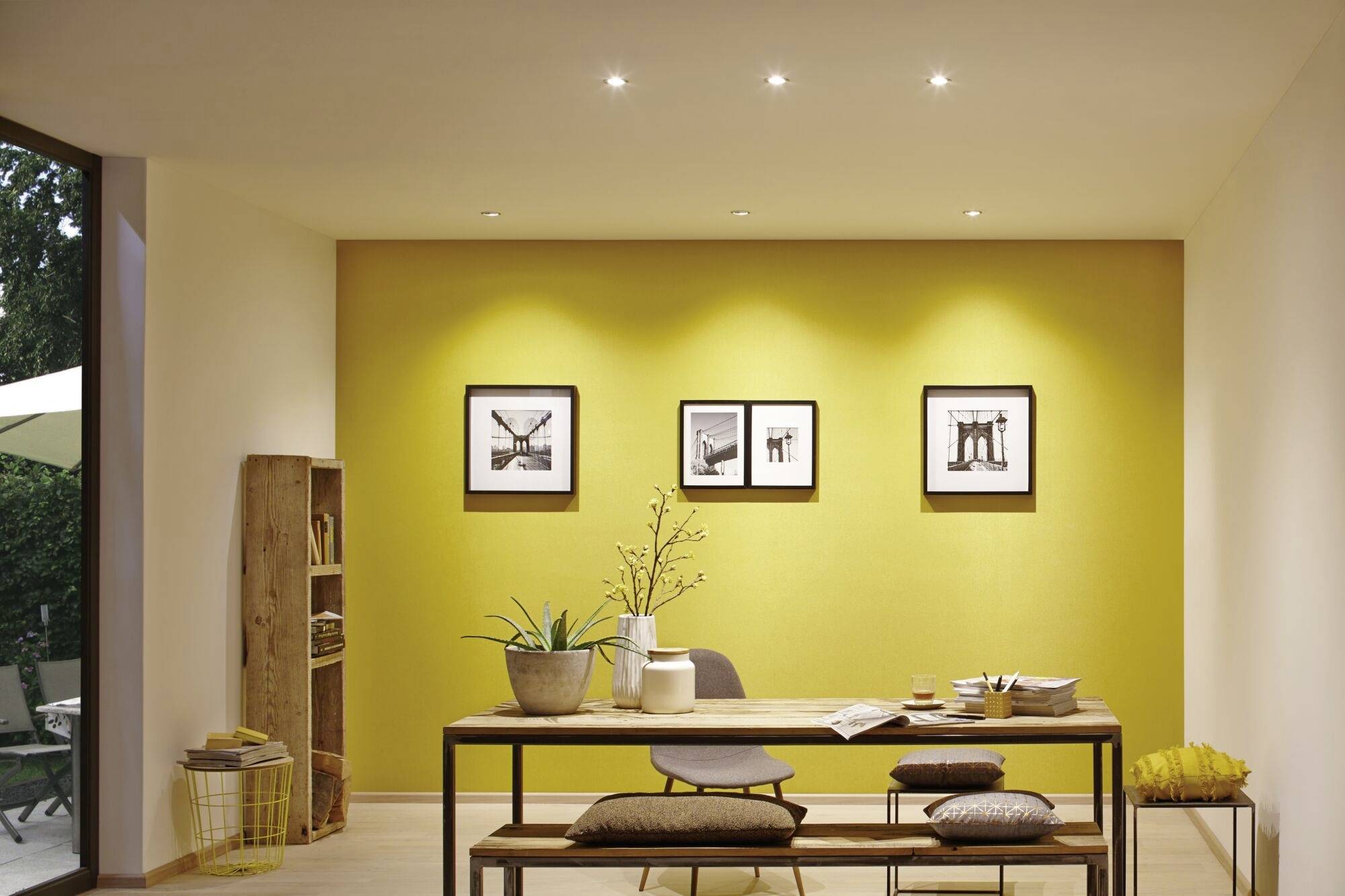 Une salle à manger avec une table en bois, deux bancs et une plante sur la table. Un mur jaune en arrière-plan avec trois photos encadrées.