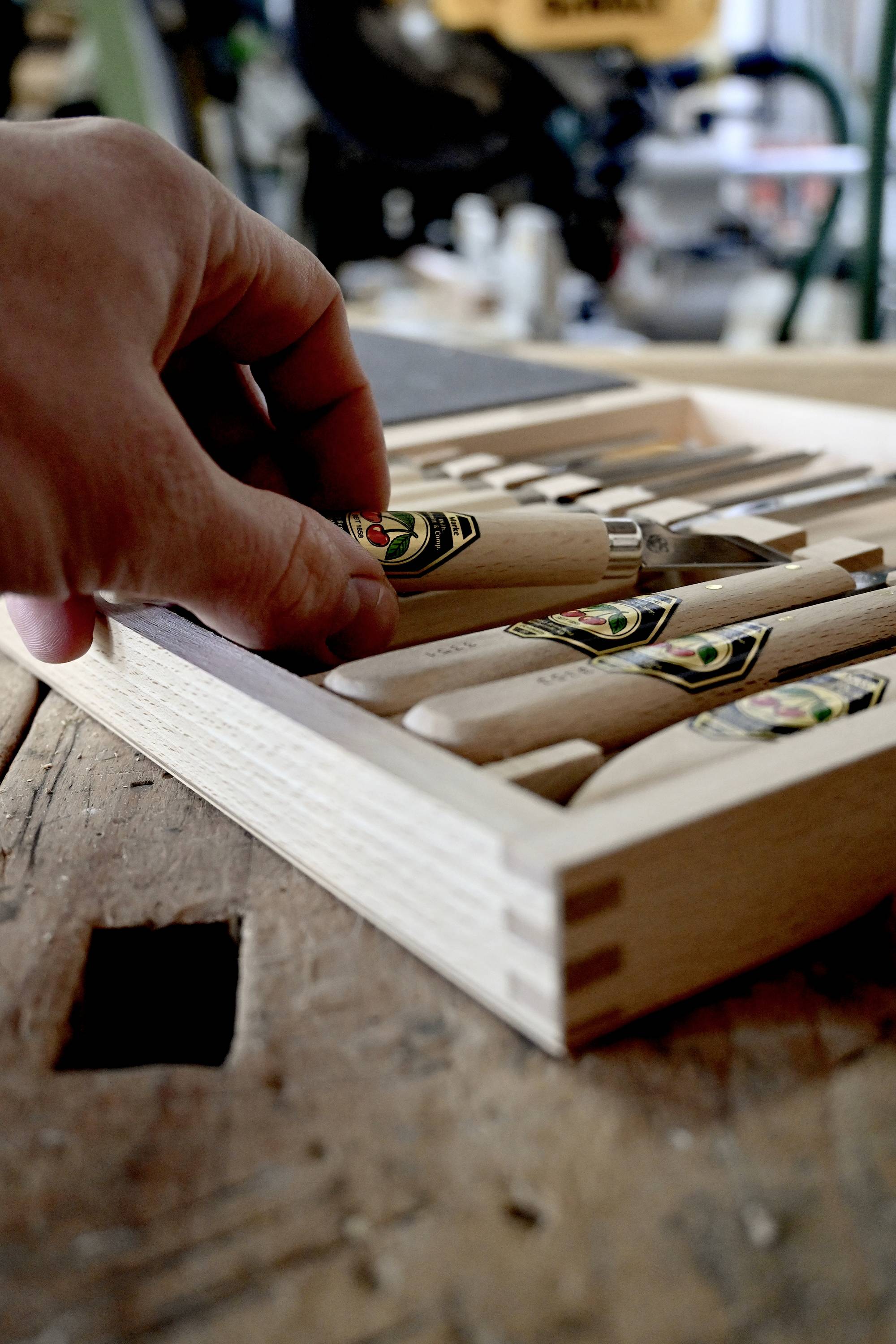 Une main saisit un outil de sculpture sur bois d'une boîte en bois posée sur un établi. À l'arrière-plan, des outils d'atelier sont flous.