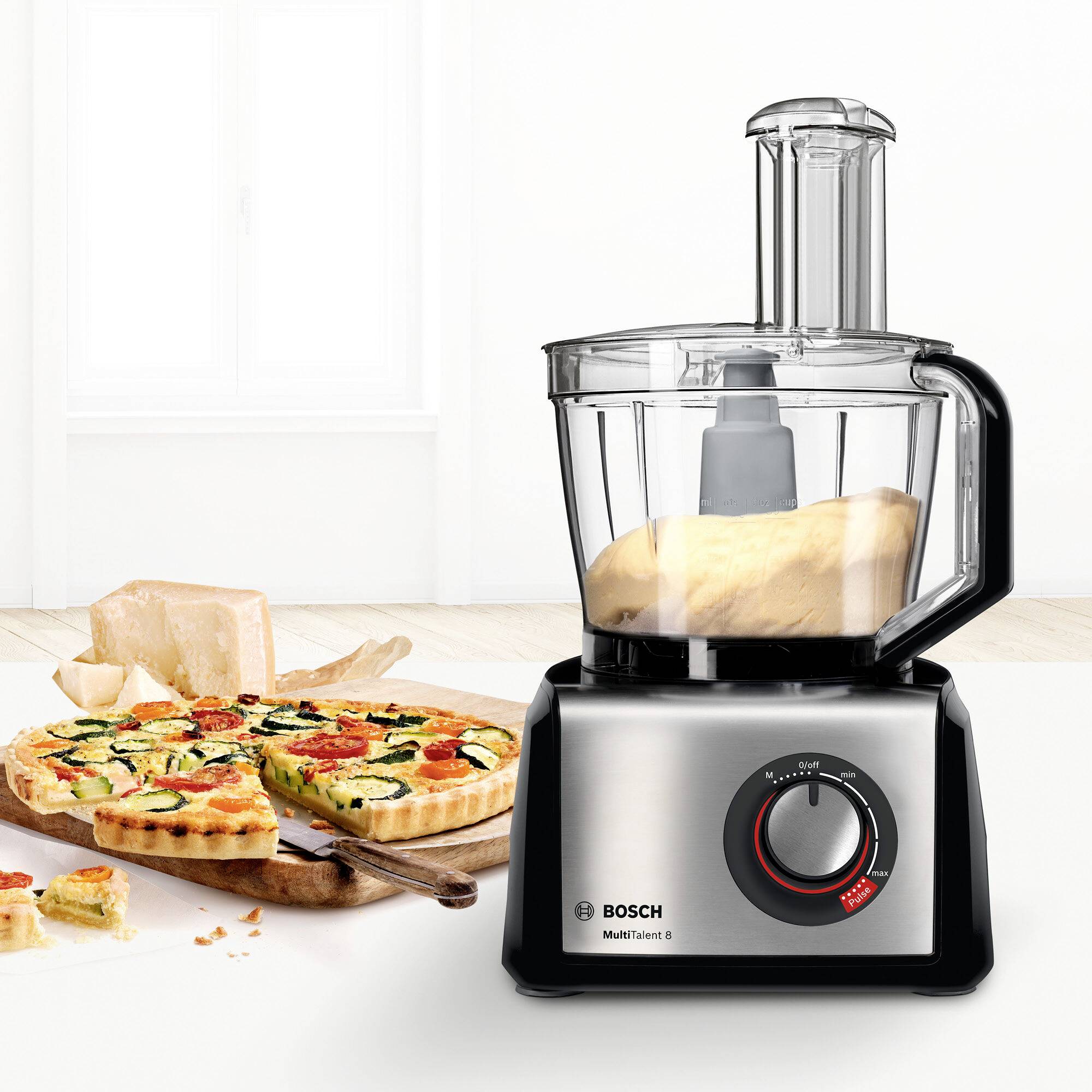 Robot de cuisine sur le plan de travail à côté du pain et de la pizza tranchés. Marque Bosch, modèle MUM5, avec mixeur intégré.