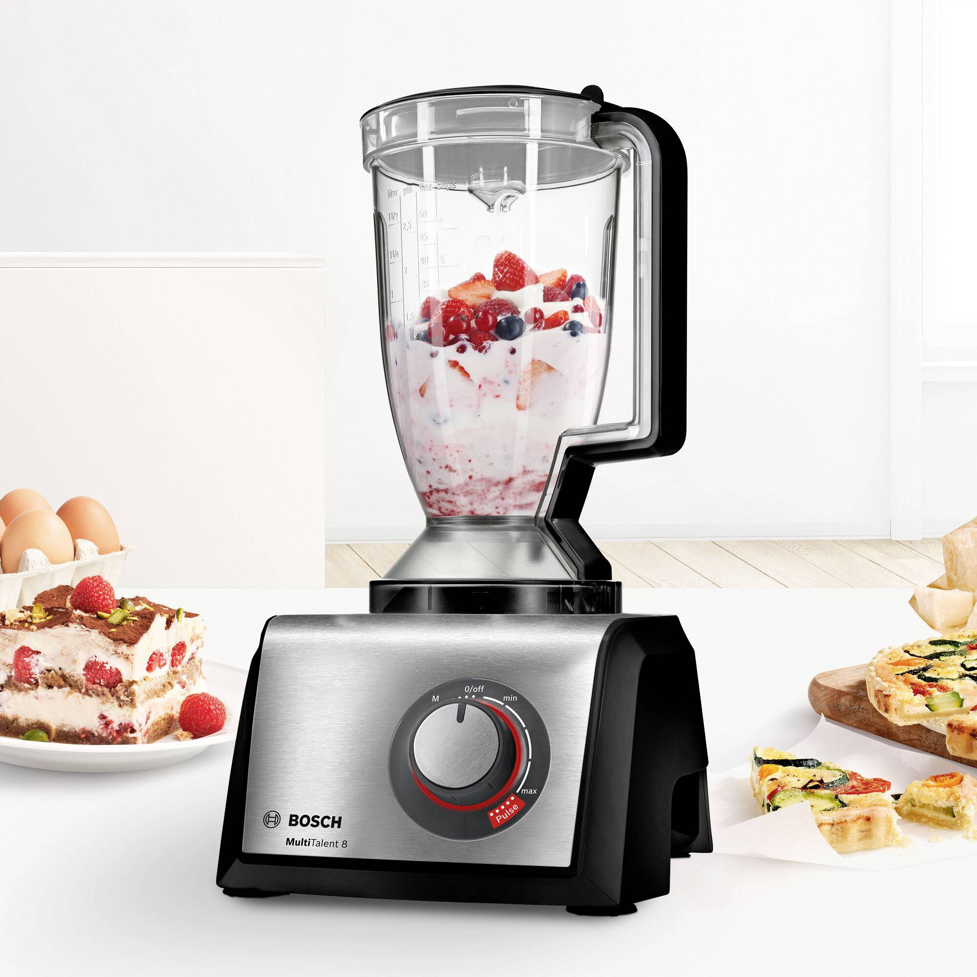 Un mixeur de Bosch est posé sur un plan de travail de cuisine, entouré de fruits et de gâteaux. Le mixeur contient un mélange de smoothie.