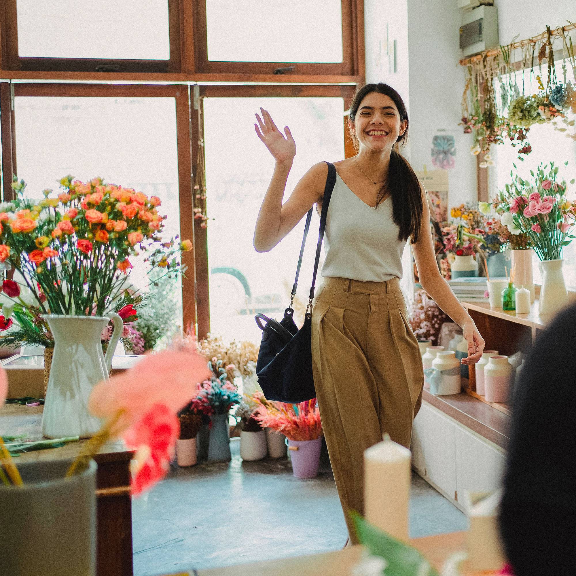 Une femme dans une boutique de fleurs, souriante et faisant un signe de la main, entourée de fleurs et de vases posés sur des tables.