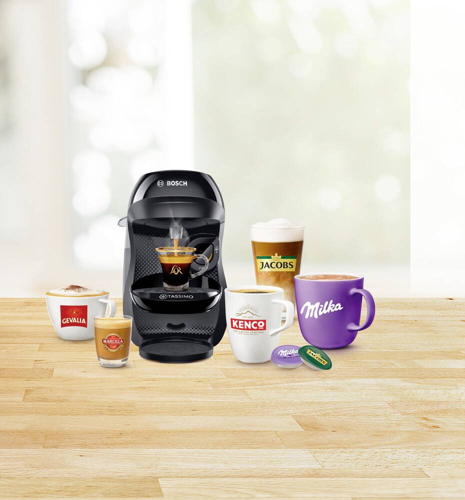 Machine à café à capsules Bosch sur une table en bois avec différentes tasses et capsules de café de marques, dont Jacobs, Milka, Gevalia et Kenco.