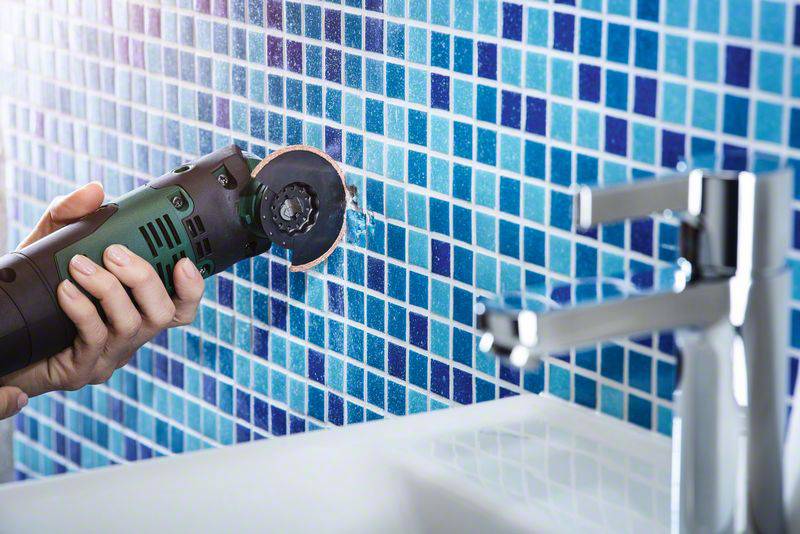Une personne coupe une mosaïque de carrelage bleu avec un meuleuse d'angle électrique dans une salle de bain.