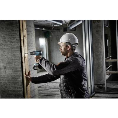 Un ouvrier portant un casque et des vêtements de travail utilise une visseuse sans fil pour percer une vis dans un mur en bois, dans un bâtiment en construction.