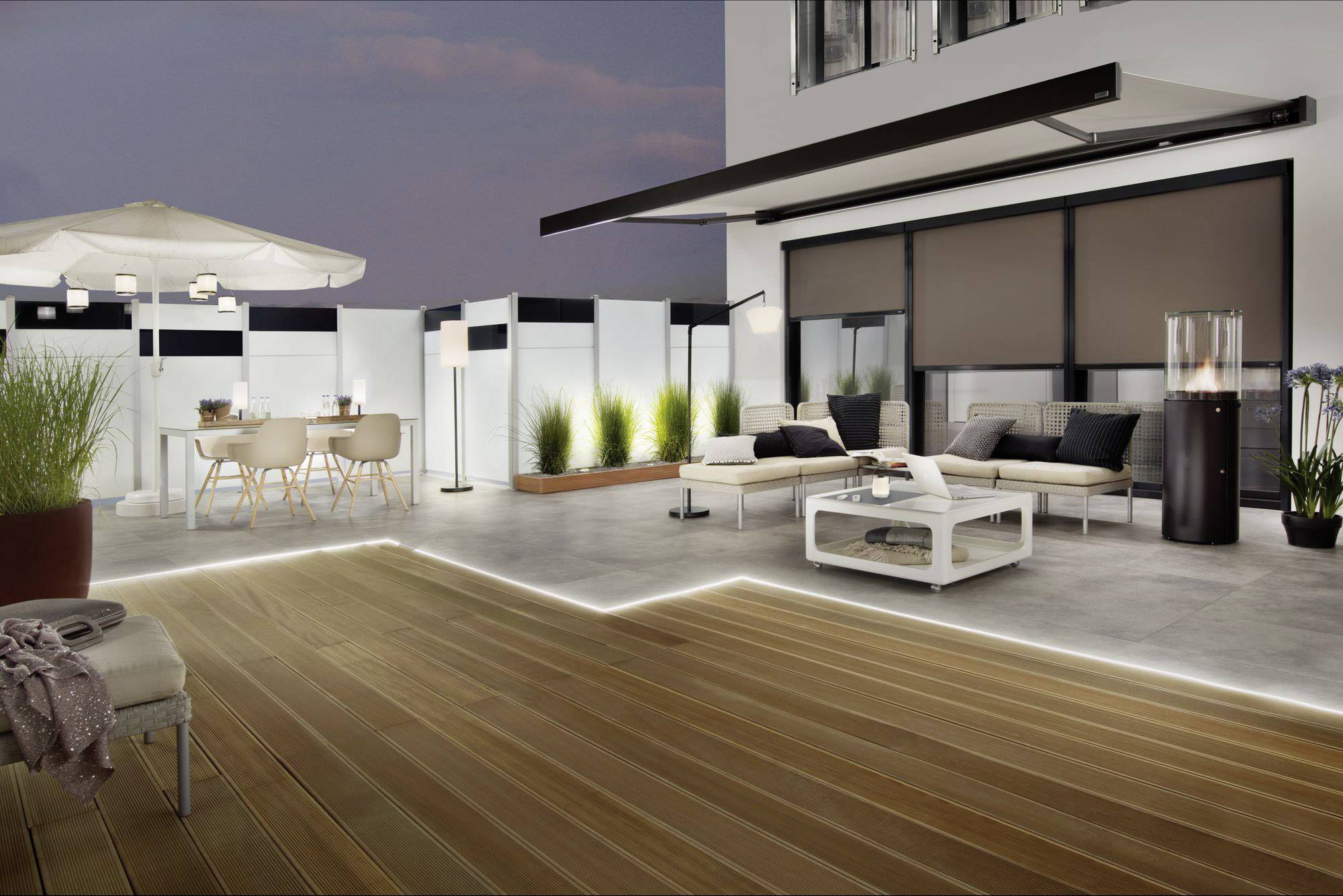 Terrasse moderne au crépuscule avec espace de détente, table à manger, parasol et arrangements de plantes, éclairée par des bandes LED.