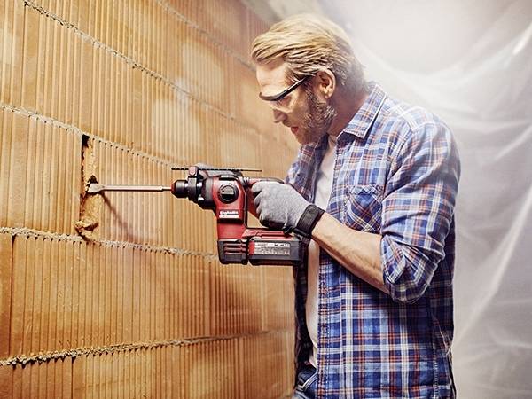 Un homme portant des lunettes de protection et des gants fore un trou dans un mur de briques à l'aide d'une perceuse rouge.