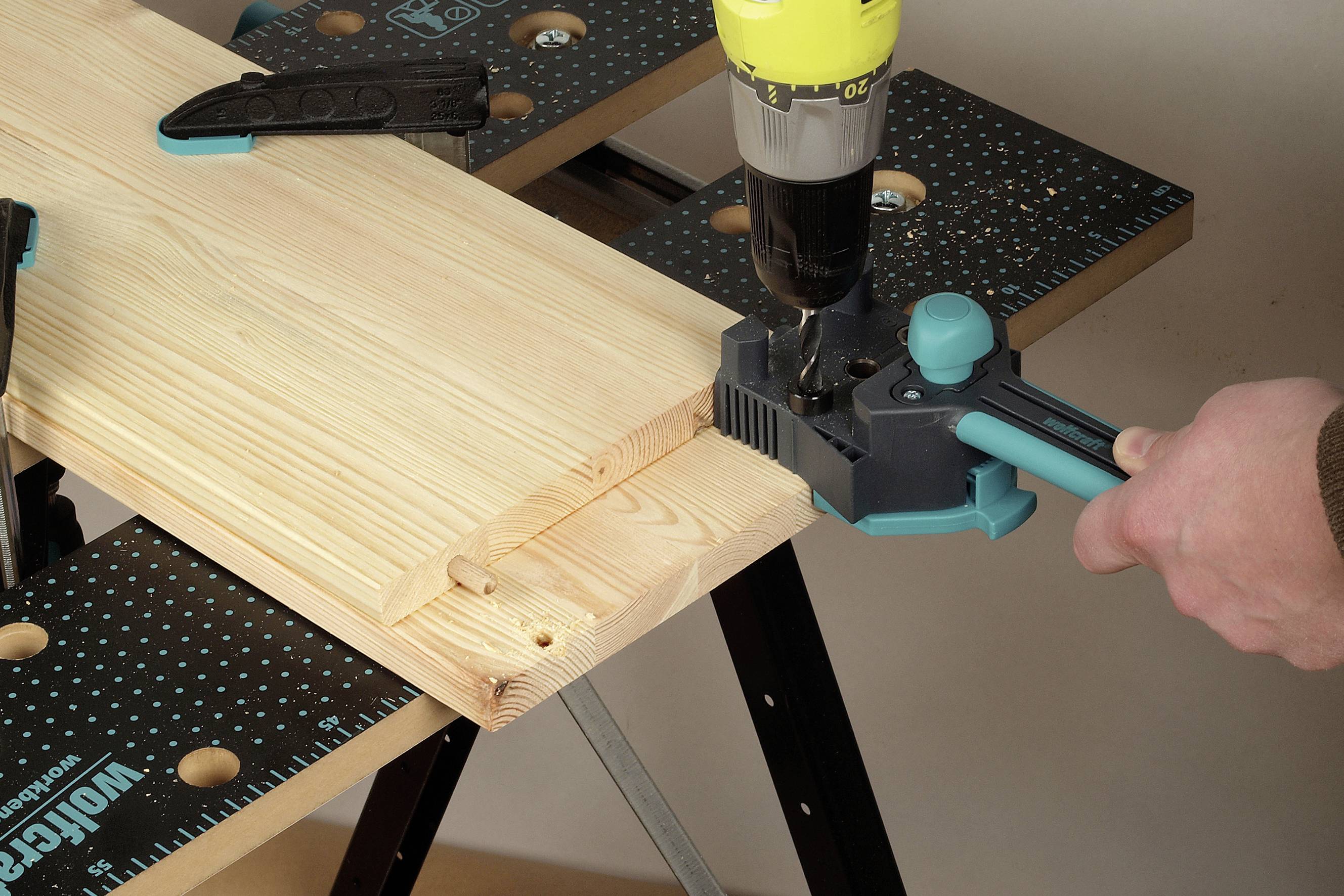 Une main fore un trou dans une planche de bois fixée sur une table de travail à l'aide d'une perceuse.