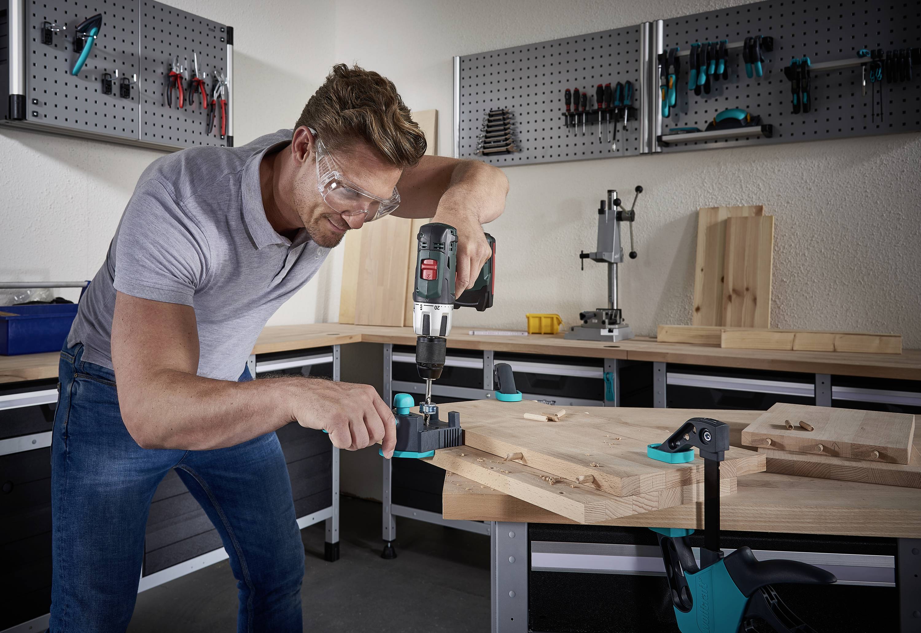 Un homme portant des lunettes de protection utilise une perceuse pour travailler le bois sur un établi. Des outils sont accrochés au mur en arrière-plan.