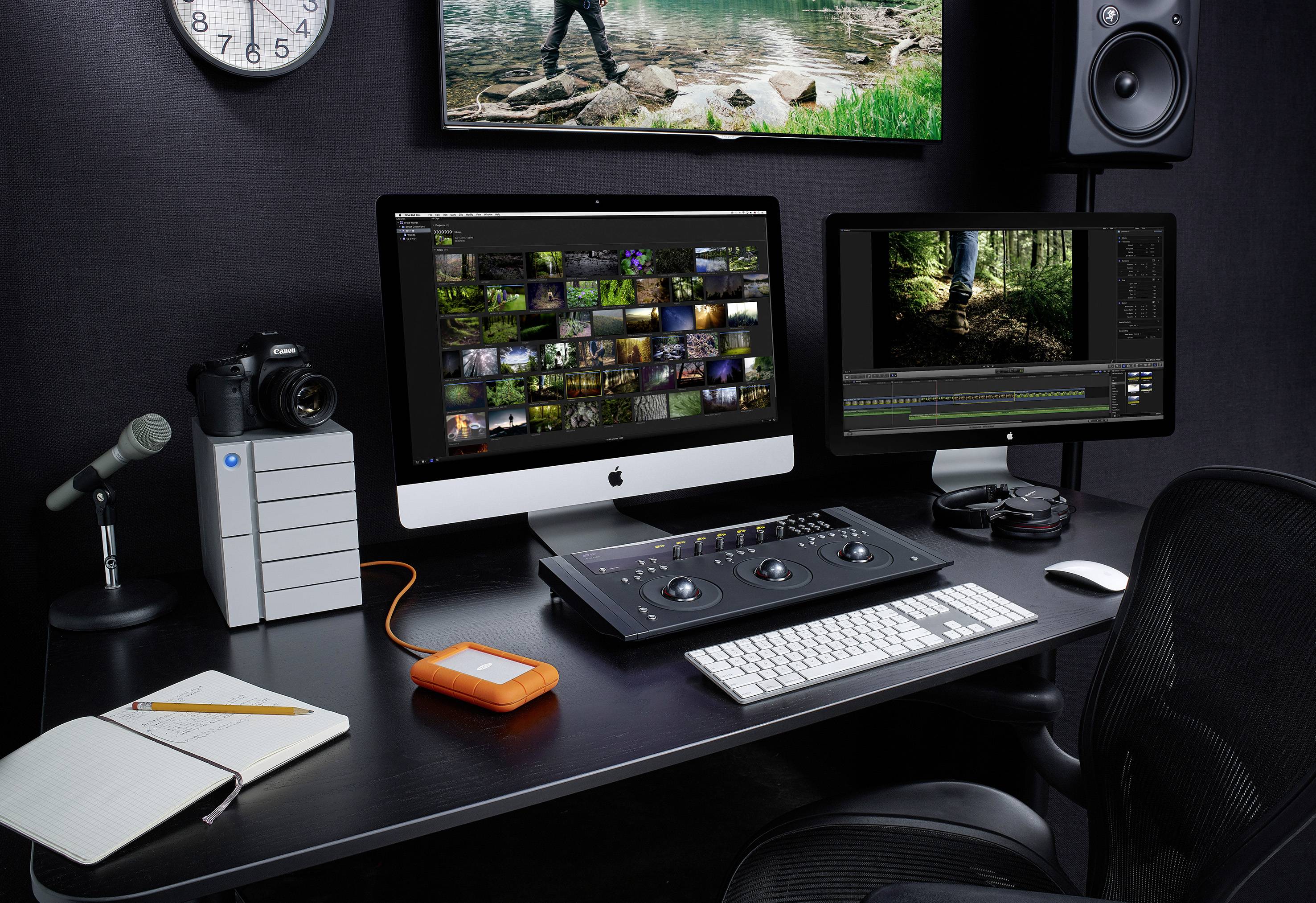 Poste de travail avec deux écrans, une console de mixage audio, une caméra, un microphone, un carnet de notes et un disque dur externe, adapté au montage multimédia.