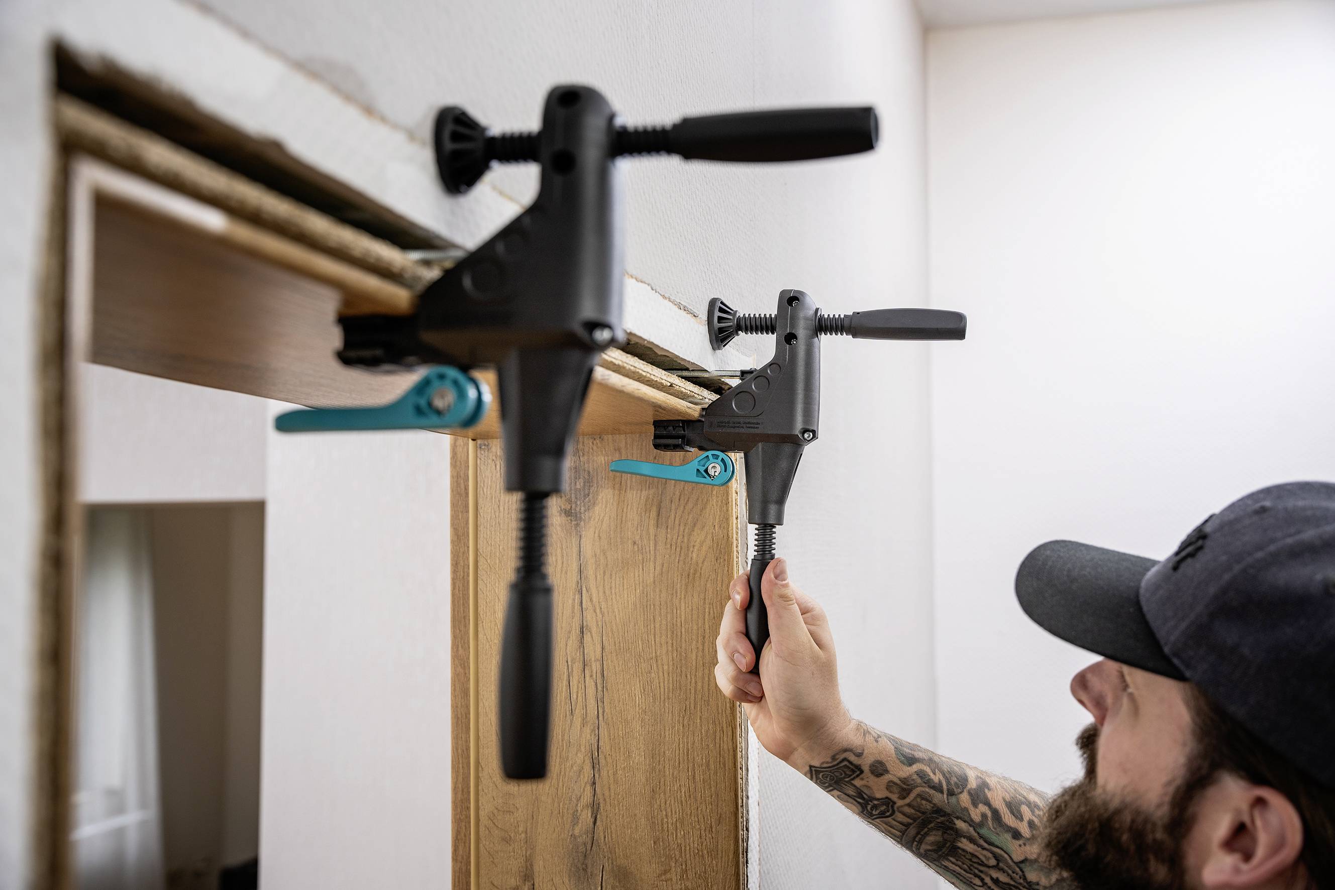 Une personne fixe une planche en bois sur un chambranle à l'aide d'un serre-joint, probablement lors de travaux de rénovation.