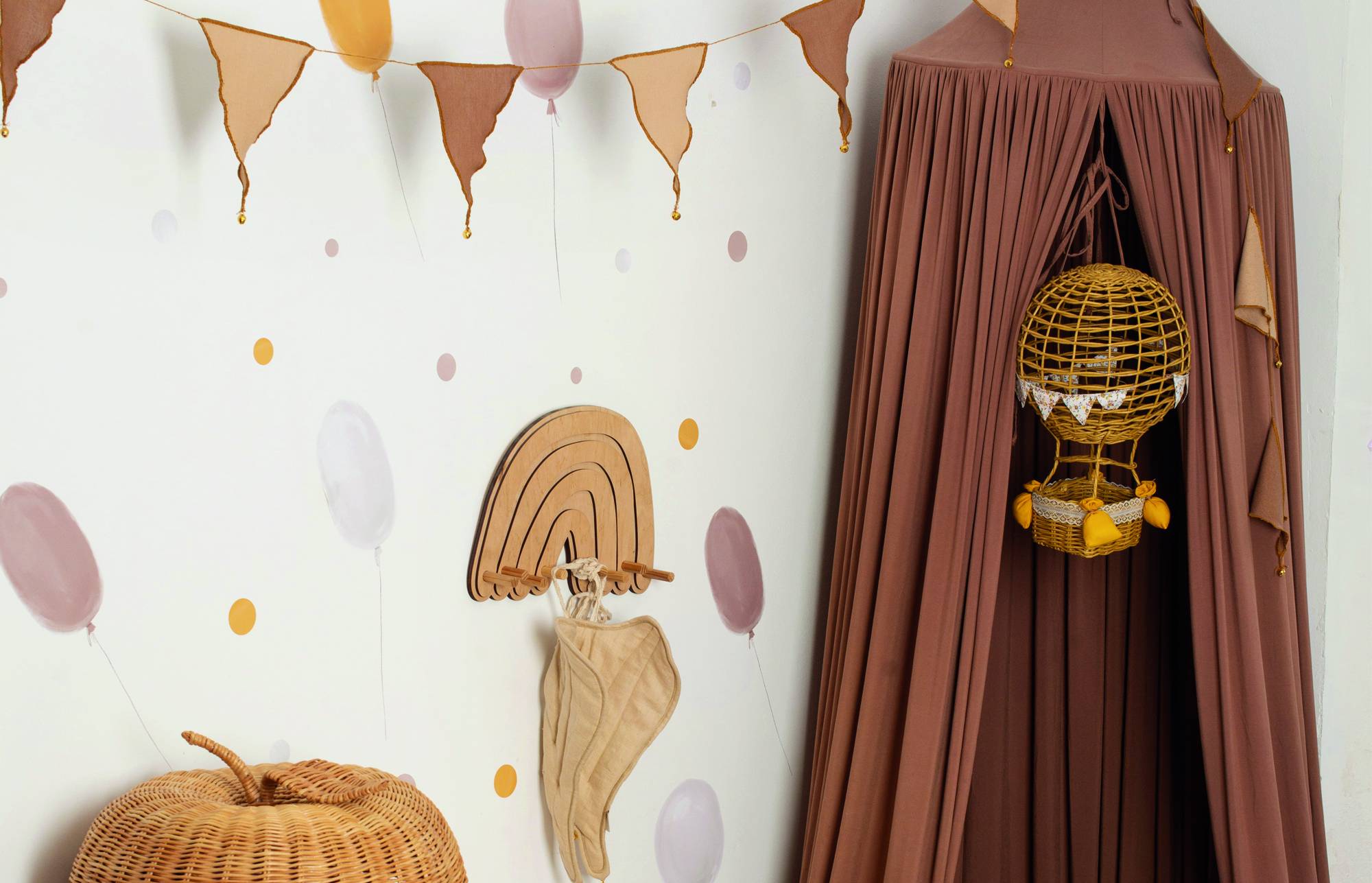 Coin de pépinière avec un dais marron et un jouet panier montgolfière suspendu à côté d'un mur décoré de ballons pastel, de guirlandes et d'un crochet arc-en-ciel.