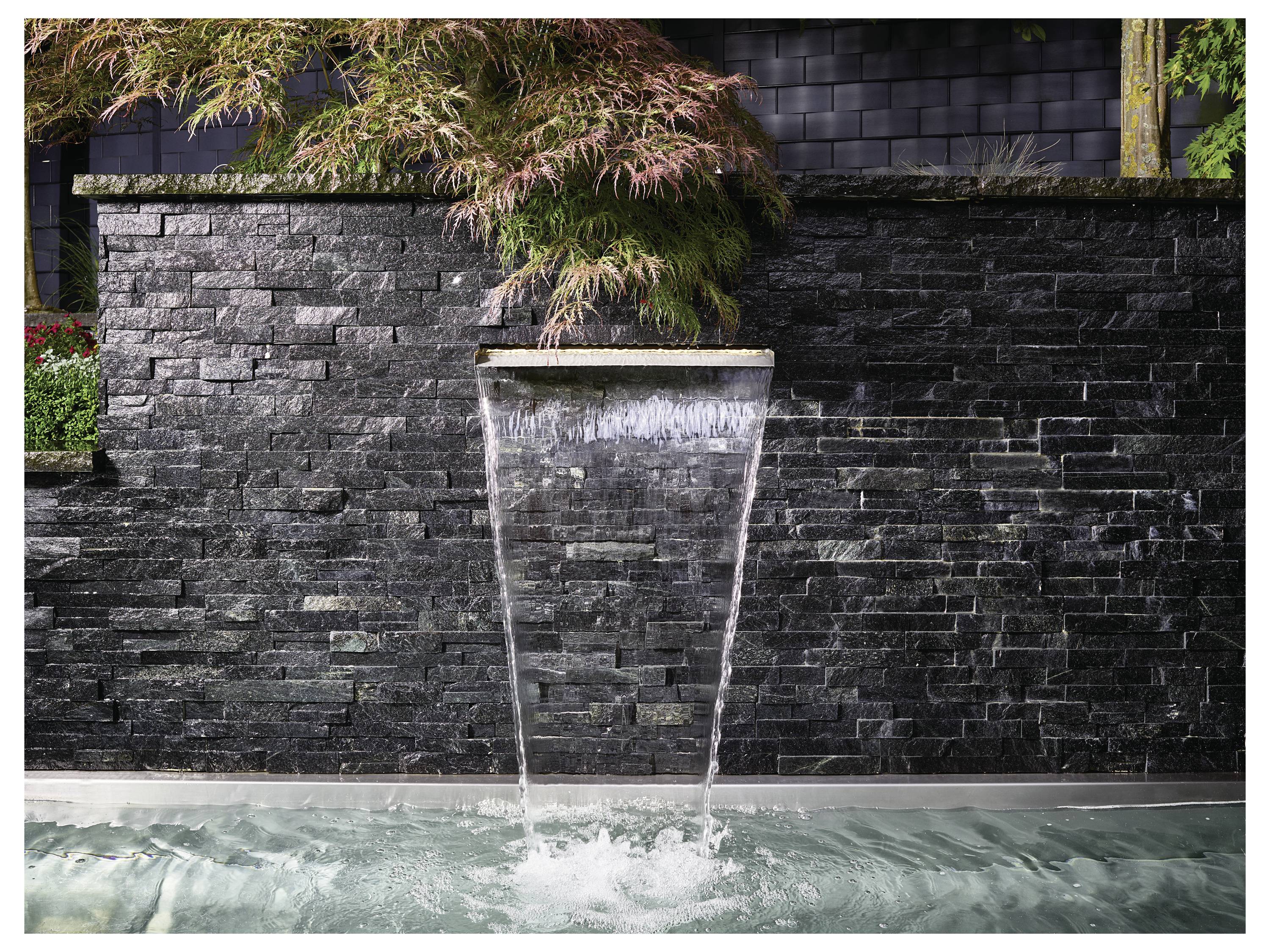 Un élément de cascade moderne coule d'un mur de pierre vers un bassin tranquille, entouré de verdure luxuriante, créant une ambiance sereine.