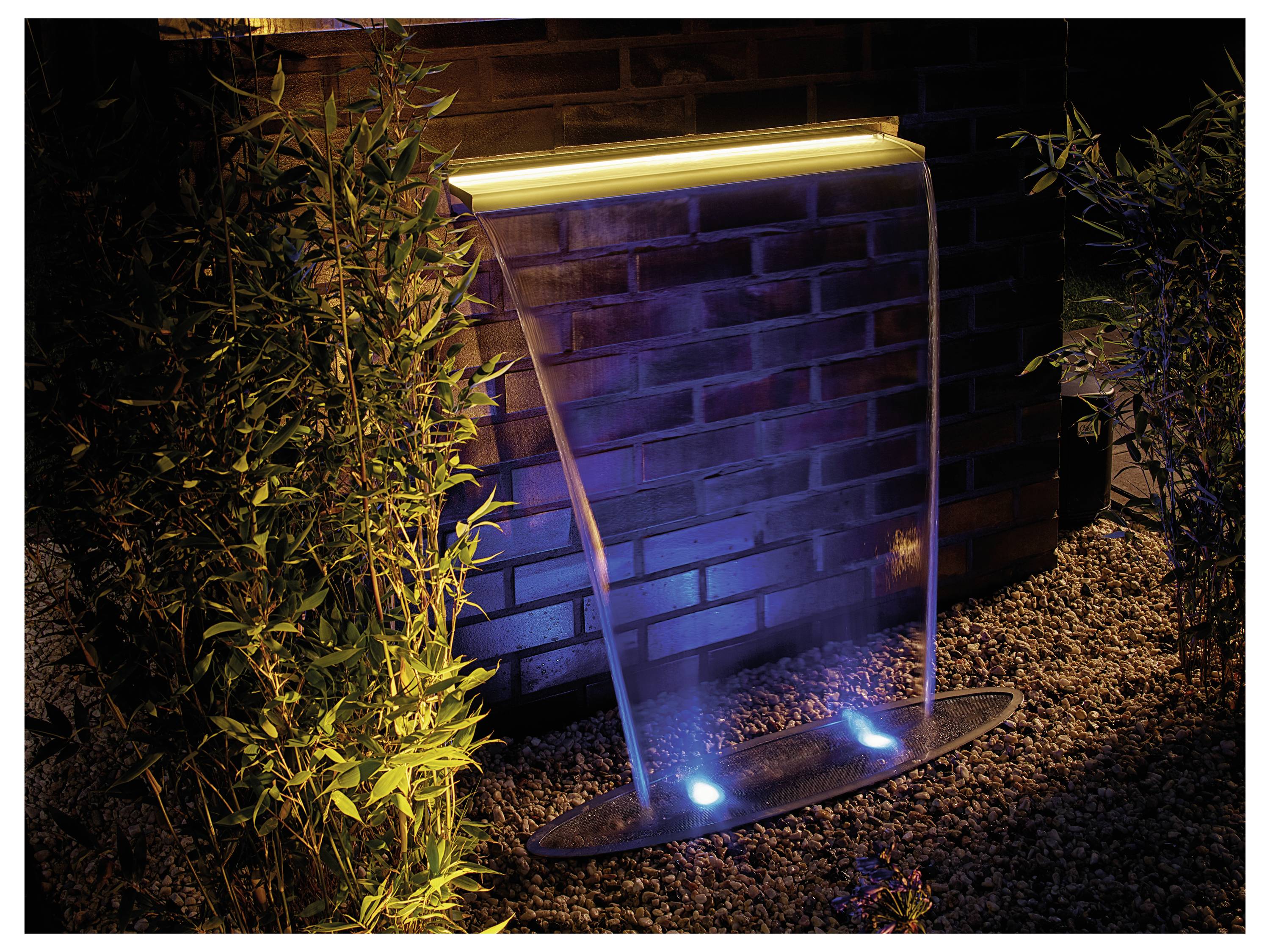 «Scène nocturne d'une fontaine moderne adossée à un mur de briques, avec des jets d'eau illuminés et des lumières bleues sur un lit de galets.»