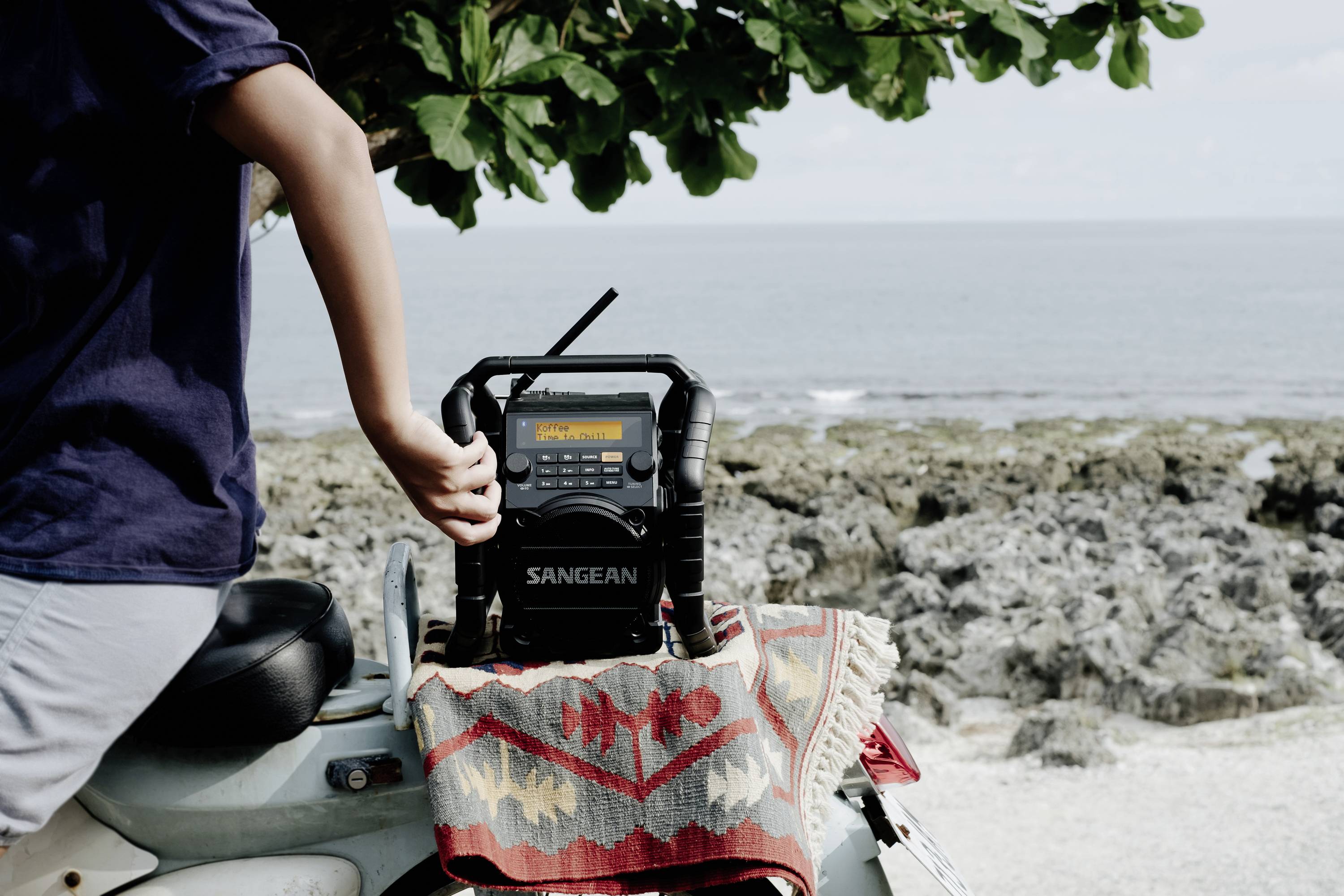 Une personne se tient à côté d'une section de côte et touche une radio portable posée sur un scooter. La mer et des rochers sont visibles en arrière-plan.