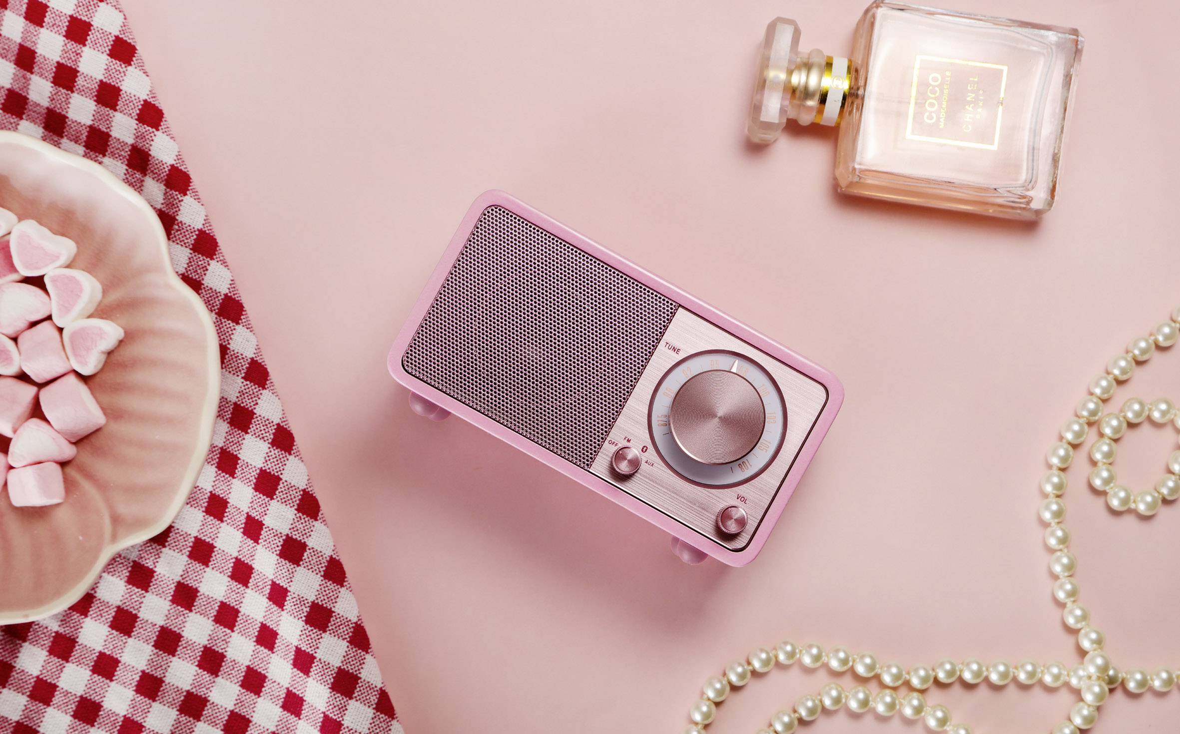 Un poste de radio rétro rose, un flacon de parfum, des bonbons en forme de cœur sur une assiette et un collier de perles sont posés sur un fond rose.