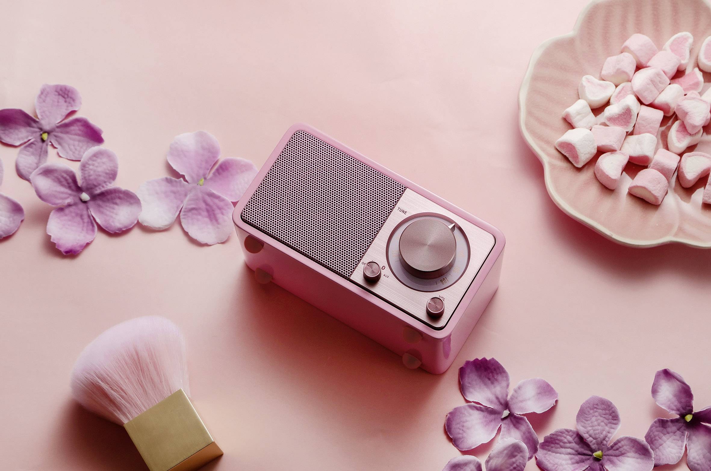 Petit poste de radio rose sur un fond rose parsemé de fleurs et de friandises. Thème principal : design rétro et esthétique pastel.