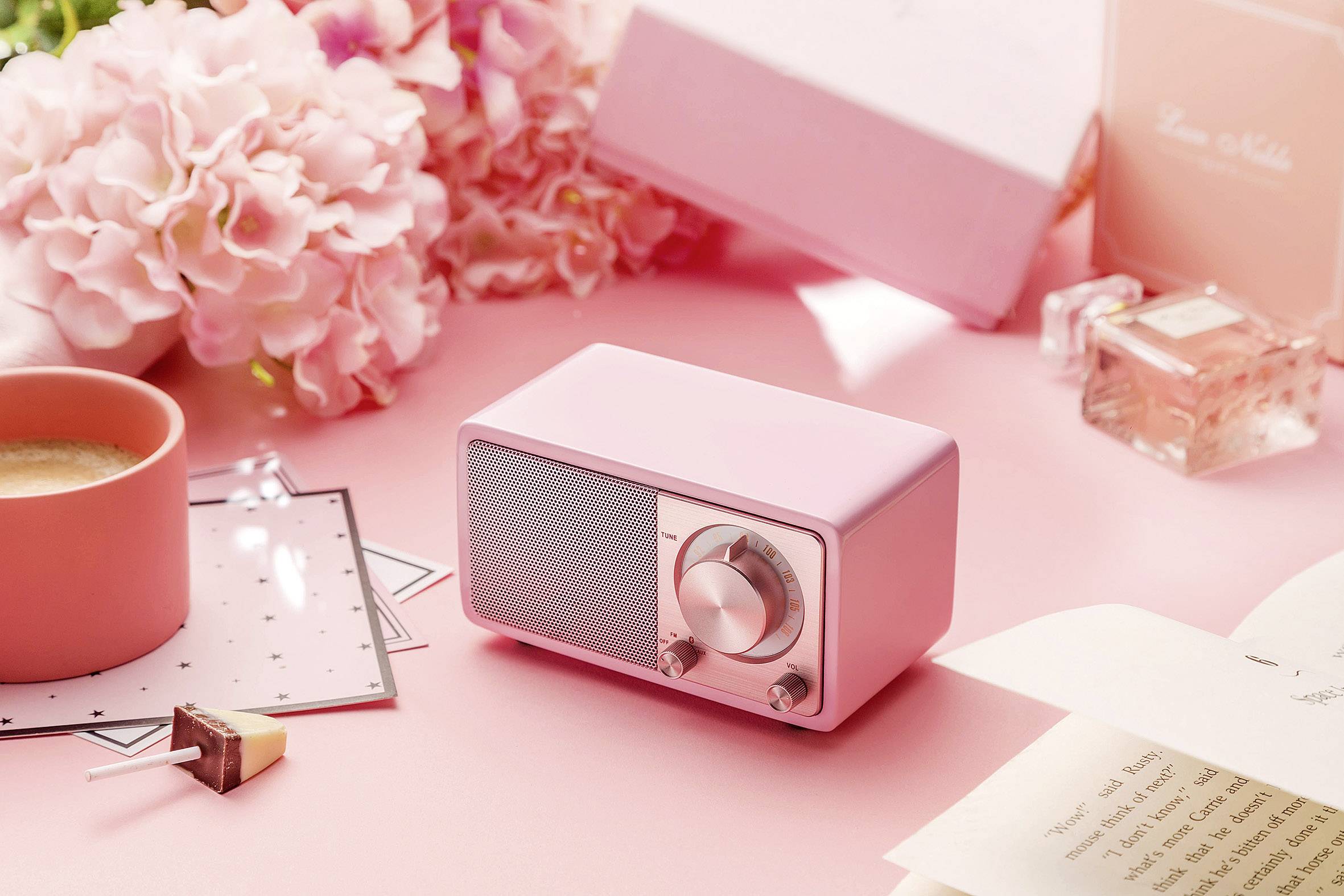 Une petite radio rose est posée sur une table, entourée de fleurs, d'une tasse de café et d'un livre ouvert.