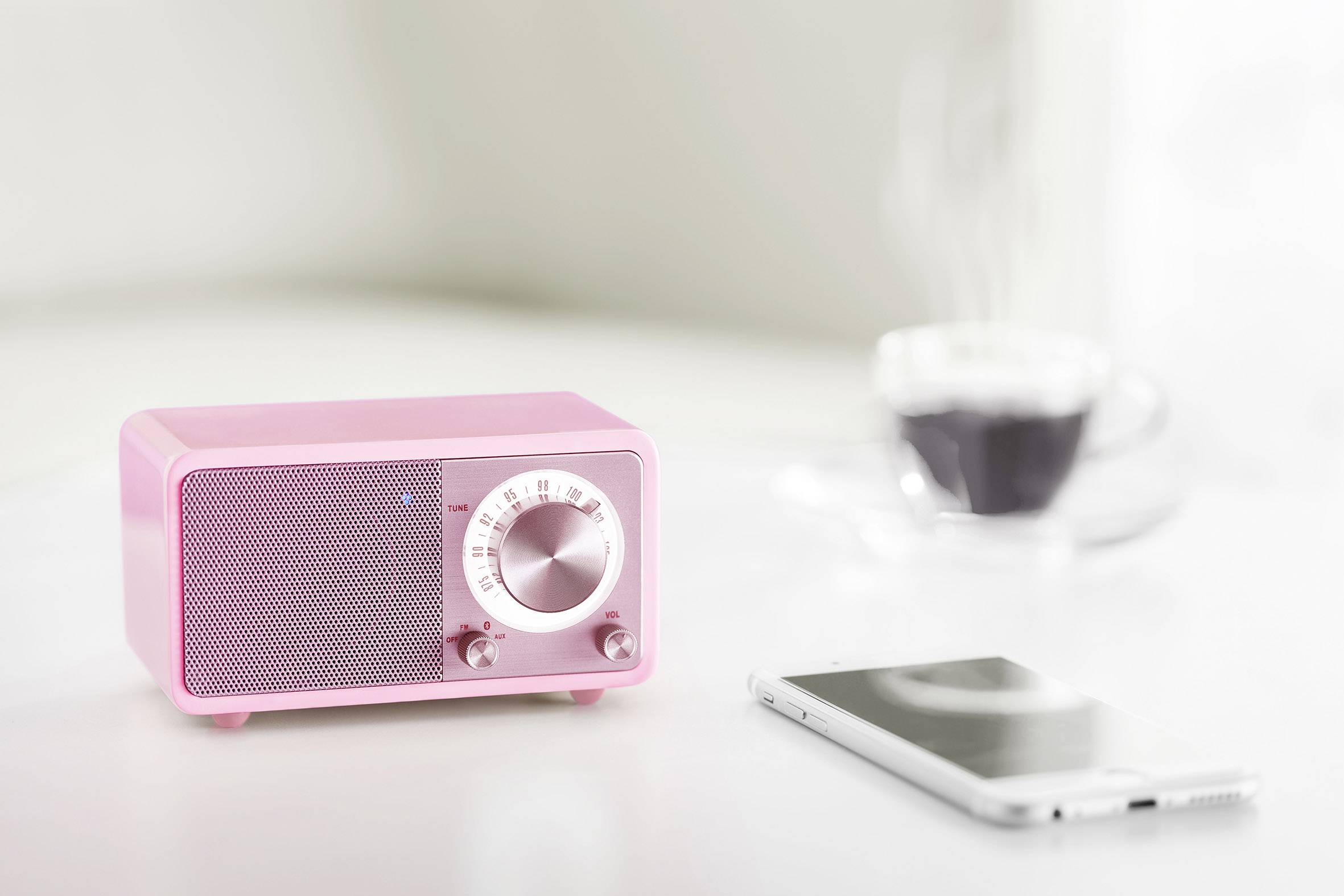 Un poste de radio rétro rose est posé sur une table, à côté d'un smartphone et d'une tasse de café fumante en arrière-plan.