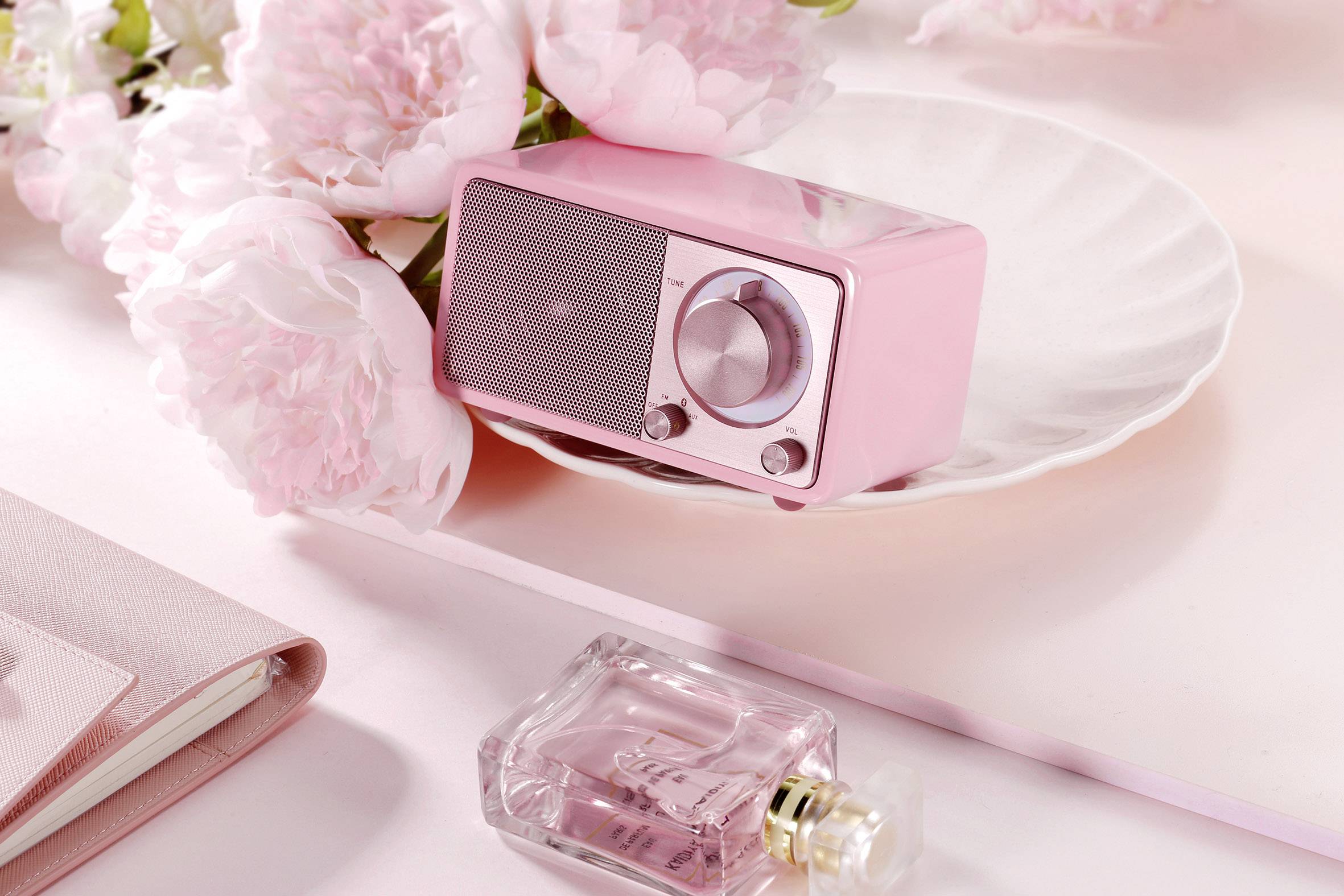 Un poste de radio rétro rose est posé sur une assiette, entouré de pivoines roses. Un flacon de parfum est posé à côté sur la table.