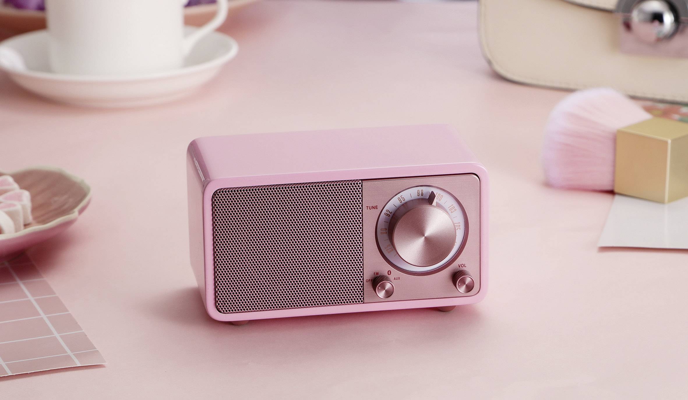 Un poste de radio rétro rose, avec sa grille de haut-parleur et ses boutons rotatifs, est posé sur une table rose. À l'arrière-plan, on aperçoit des tasses et des pinceaux.