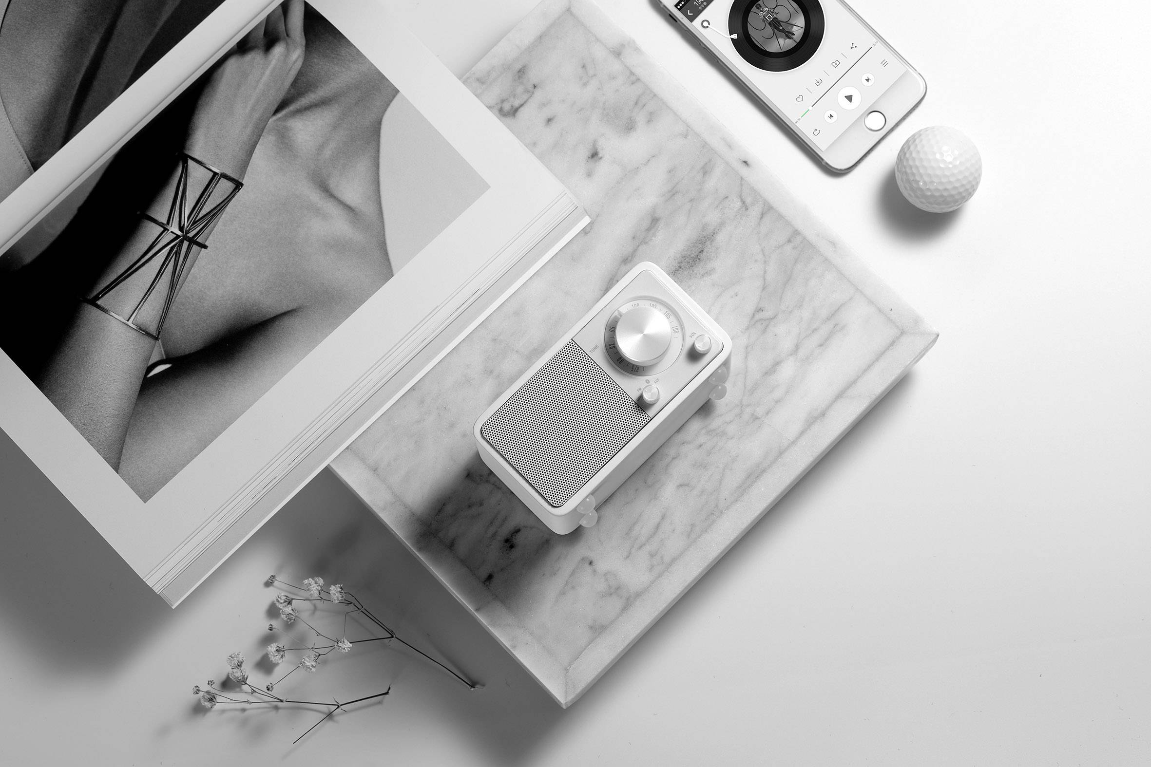 Un livre de mode ouvert, une radio posée sur une table en marbre et une balle de golf reposent sur une surface blanche, baignés dans une lumière douce.