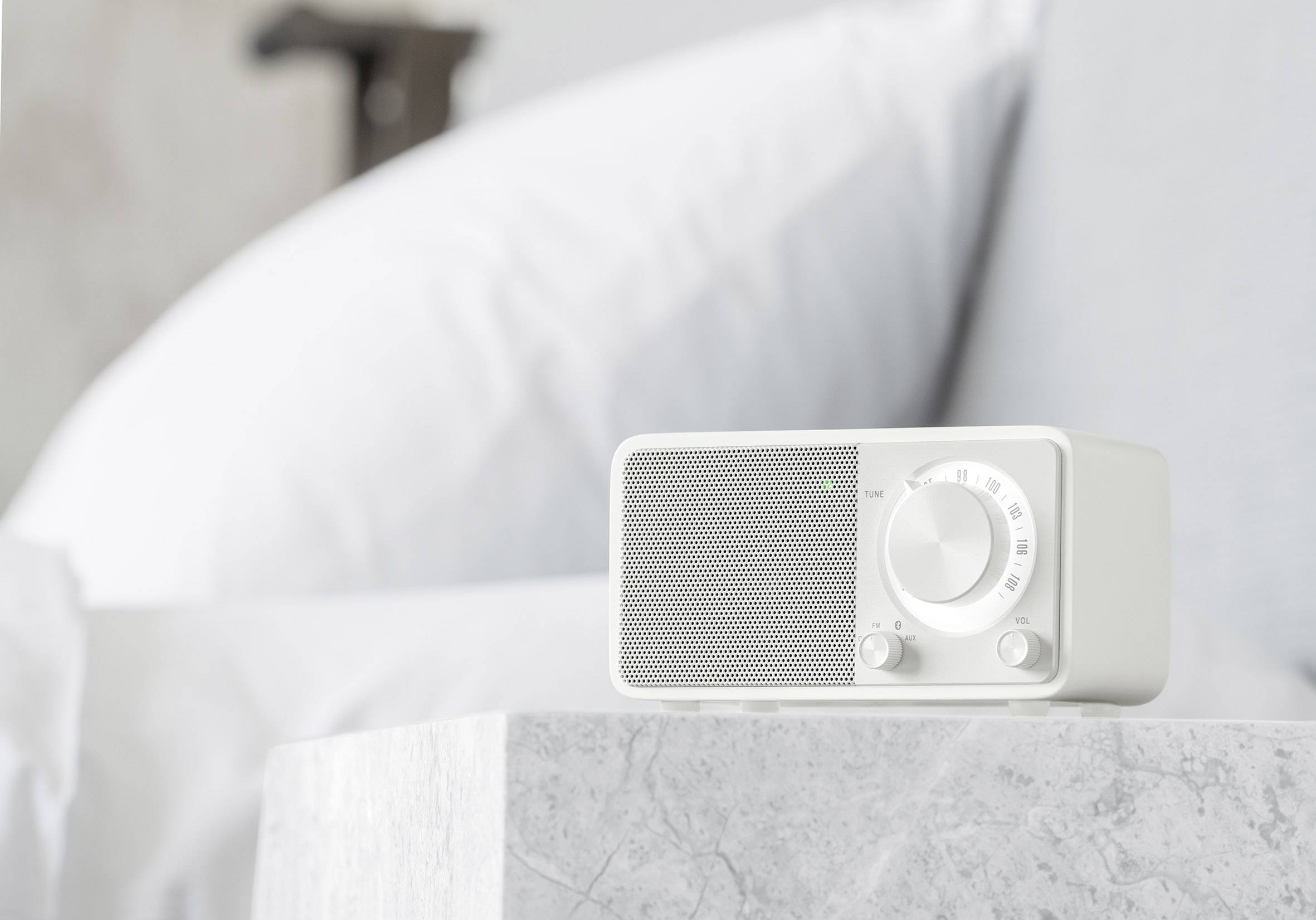 Radio portable blanc avec un régulateur de volume et un bouton de réglage, posé sur une table de nuit devant un lit aux oreillers blancs.