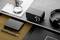 Bureau avec une loupe posée sur un journal, une radio noire, des écouteurs, un stylo et un disque dur, à côté se trouve un carnet noir.