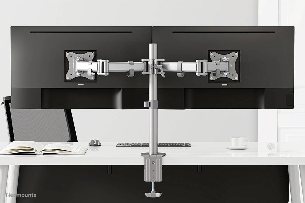 'Support de double moniteur avec deux écrans, fixé à un bureau blanc. Un livre ouvert et une tasse sont visibles en arrière-plan.'