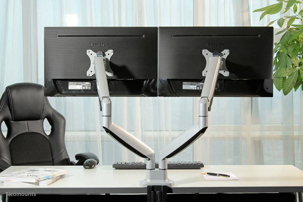 Deux écrans montés sur des bras articulés sur un bureau, avec une chaise de bureau et des plantes en arrière-plan. Un clavier, une souris et un bloc-notes sont également présents.