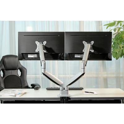 Deux écrans montés sur des bras articulés sur un bureau, avec une chaise de bureau et des plantes en arrière-plan. Un clavier, une souris et un bloc-notes sont également présents.