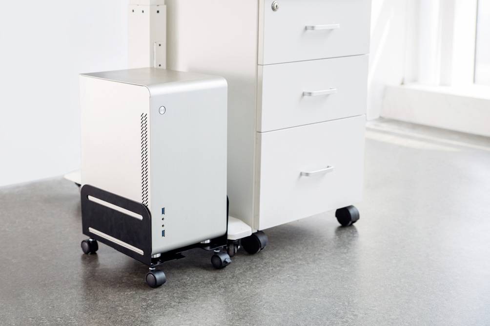 Un ordinateur blanc est posé sur un support mobile à côté d'une commode blanche avec des tiroirs, sur un sol en béton gris.