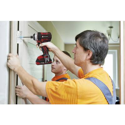 Deux hommes portant des chemises orange installent une charnière de porte à l'aide d'une visseuse sans fil. L'un maintient la porte, tandis que l'autre visse.