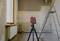 Une pièce est en rénovation, un appareil laser indique des lignes droites sur le mur, une échelle est placée à droite à côté d'un radiateur situé sous la fenêtre.