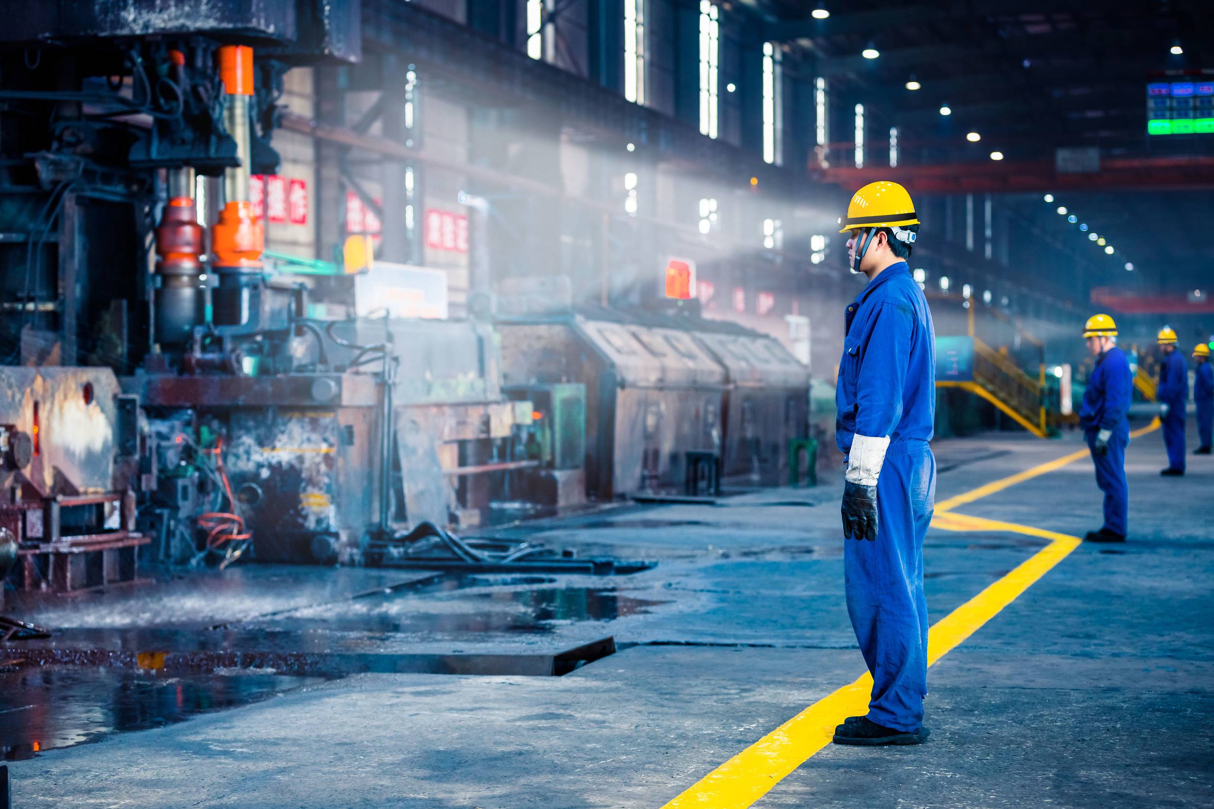 Ouvriers portant des vêtements de protection et des casques, travaillant sur une ligne de production dans un grand hall d'usine. Des machines industrielles sont visibles en arrière-plan.