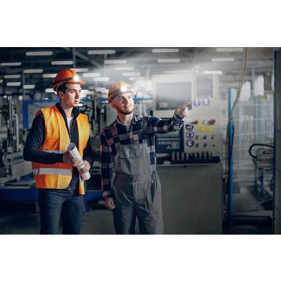Deux ouvriers portant des casques de sécurité et des gilets haute visibilité se tiennent dans un hall d'usine, l'un pointant une machine, l'autre tenant un document.
