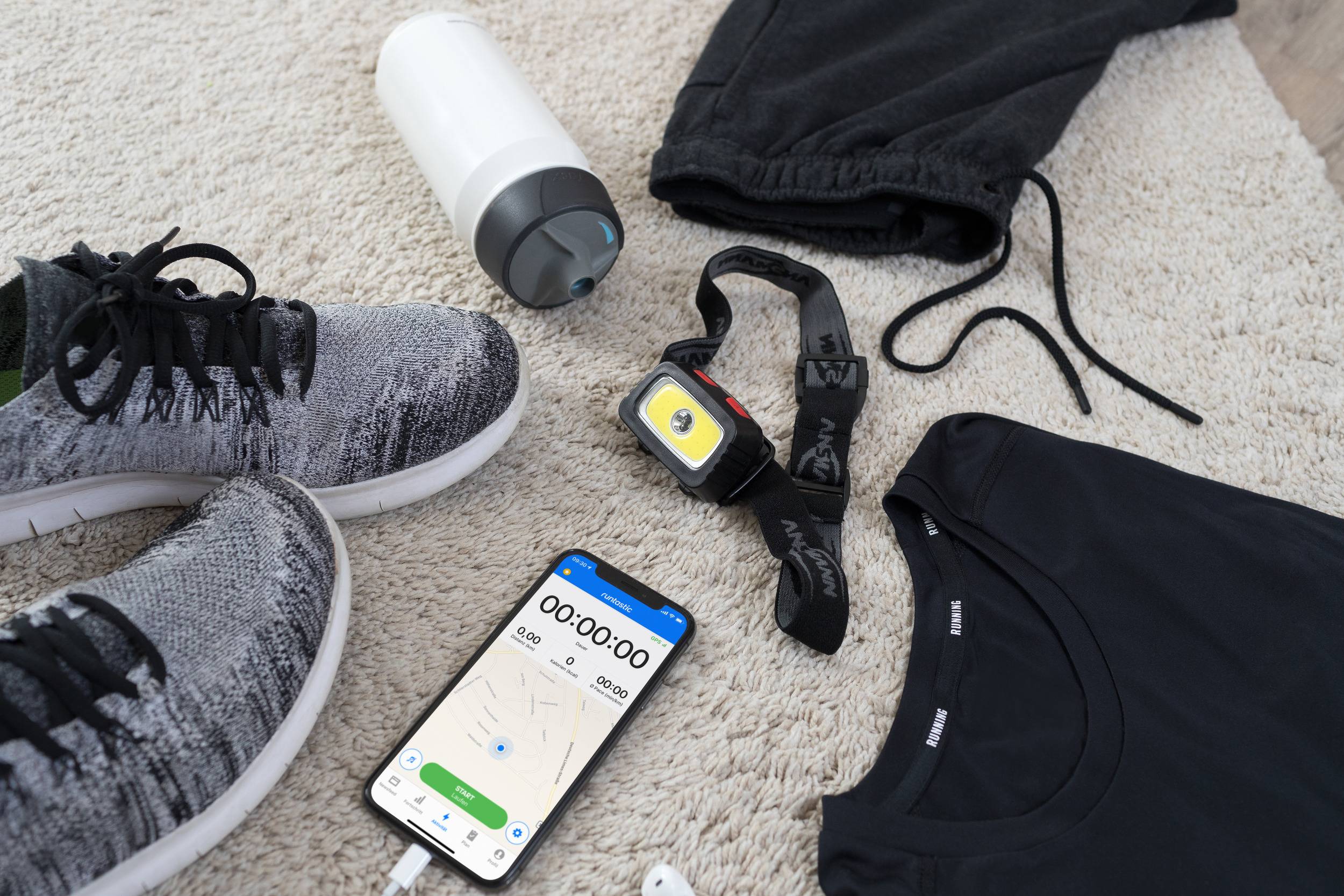Des vêtements et accessoires de sport sont disposés sur un tapis : des chaussures, une bouteille d'eau, des écouteurs, une montre de running, un pantalon de sport et un smartphone avec une application de fitness ouverte.