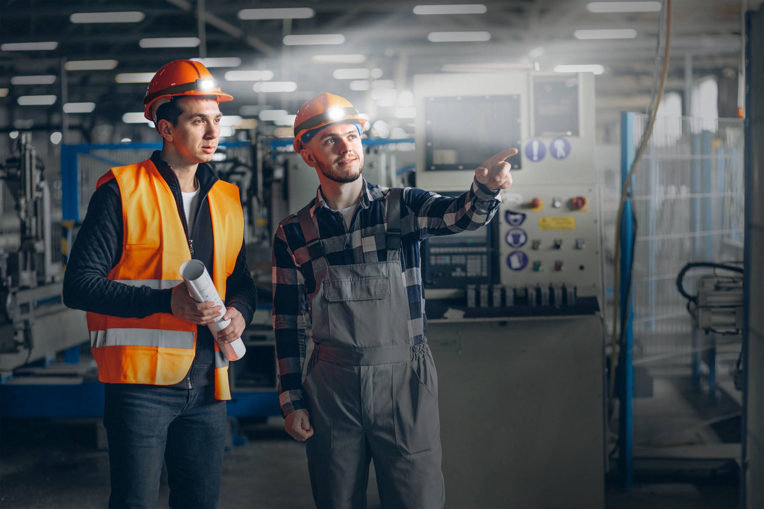 Deux ouvriers en tenue de sécurité se tiennent dans un hall d'usine. L'un tient des plans de construction, l'autre pointe une machine en arrière-plan.