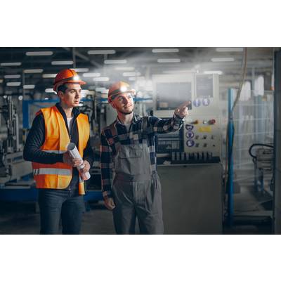 Deux ouvriers en tenue de sécurité se tiennent dans un hall d'usine. L'un tient des plans de construction, l'autre pointe une machine en arrière-plan.