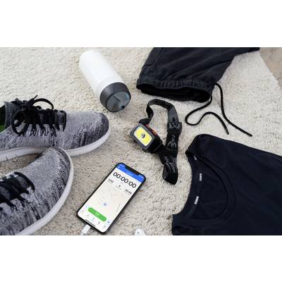 Du matériel de sport est posé sur un tapis : des chaussures de course, des vêtements de sport, une bouteille d'eau, une montre de fitness et un smartphone avec une application de chronomètre.