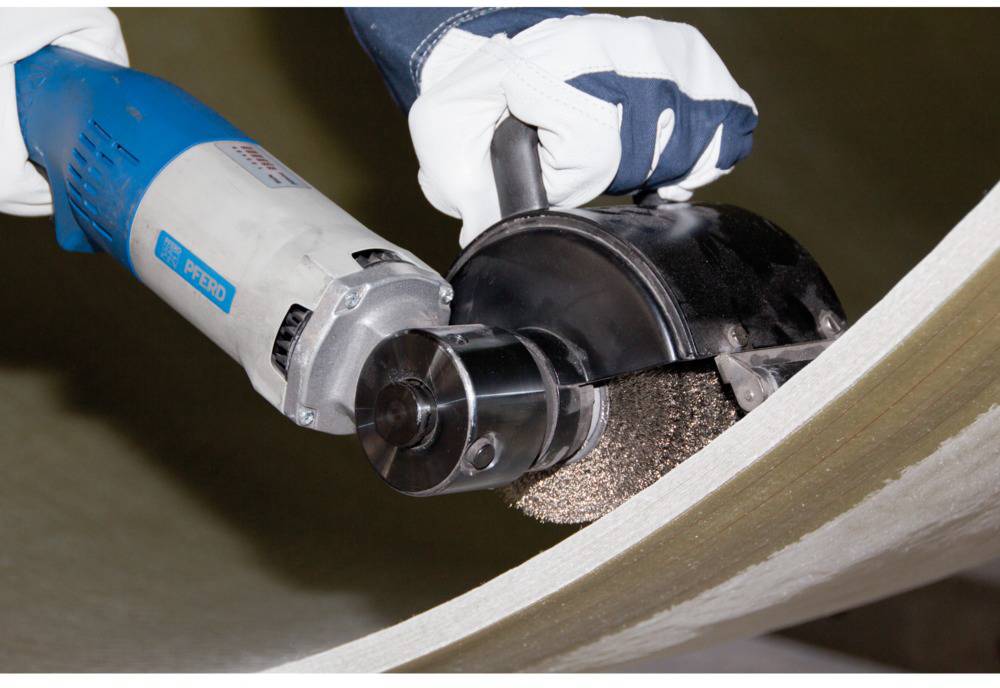 Une personne porte des gants de travail et tient un outil électrique qui ponçe ou polit une surface métallique.