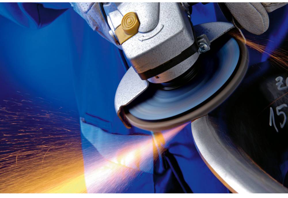 'Gros plan d'une meuleuse d'angle projetant des étincelles pendant qu'elle travaille le métal. L'arrière-plan est bleu, et une main tient l'outil.'