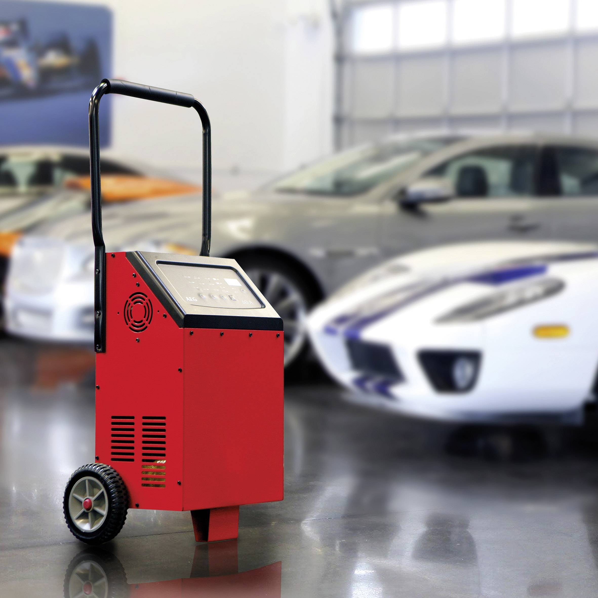 Un chargeur de batterie mobile rouge est posé dans un garage devant trois voitures. À l'arrière-plan, deux voitures de sport sont visibles de manière floue.