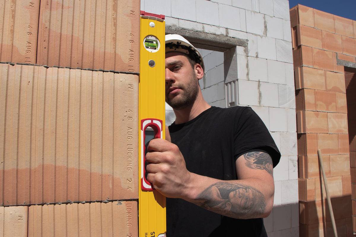 Un ouvrier utilise un niveau à bulle pour vérifier l'alignement vertical d'un mur de briques. D'autres murs sont visibles en arrière-plan.