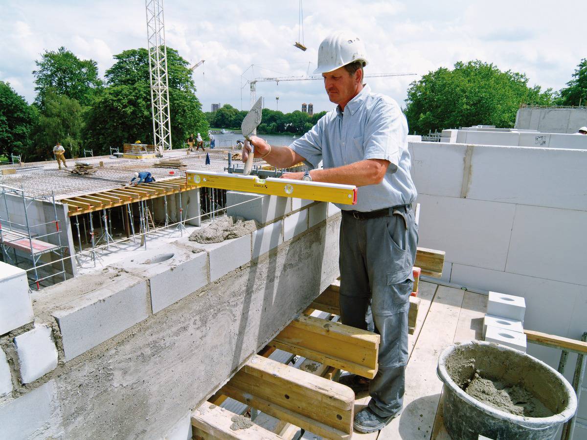 Un ouvrier portant un casque de chantier et des outils travaille sur un mur. Des grues de construction et une structure partiellement achevée sont visibles en arrière-plan.