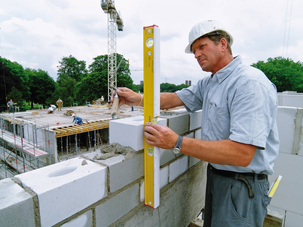 Un ouvrier vérifie la planéité d'un mur avec un niveau à bulle sur un chantier en extérieur. Un grue et d'autres travailleurs sont visibles en arrière-plan.