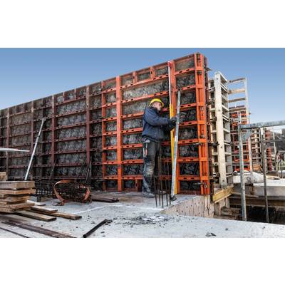 Un ouvrier portant un casque jaune ajuste des coffrages sur un chantier. Il se tient sur du béton, entouré de matériaux de construction.